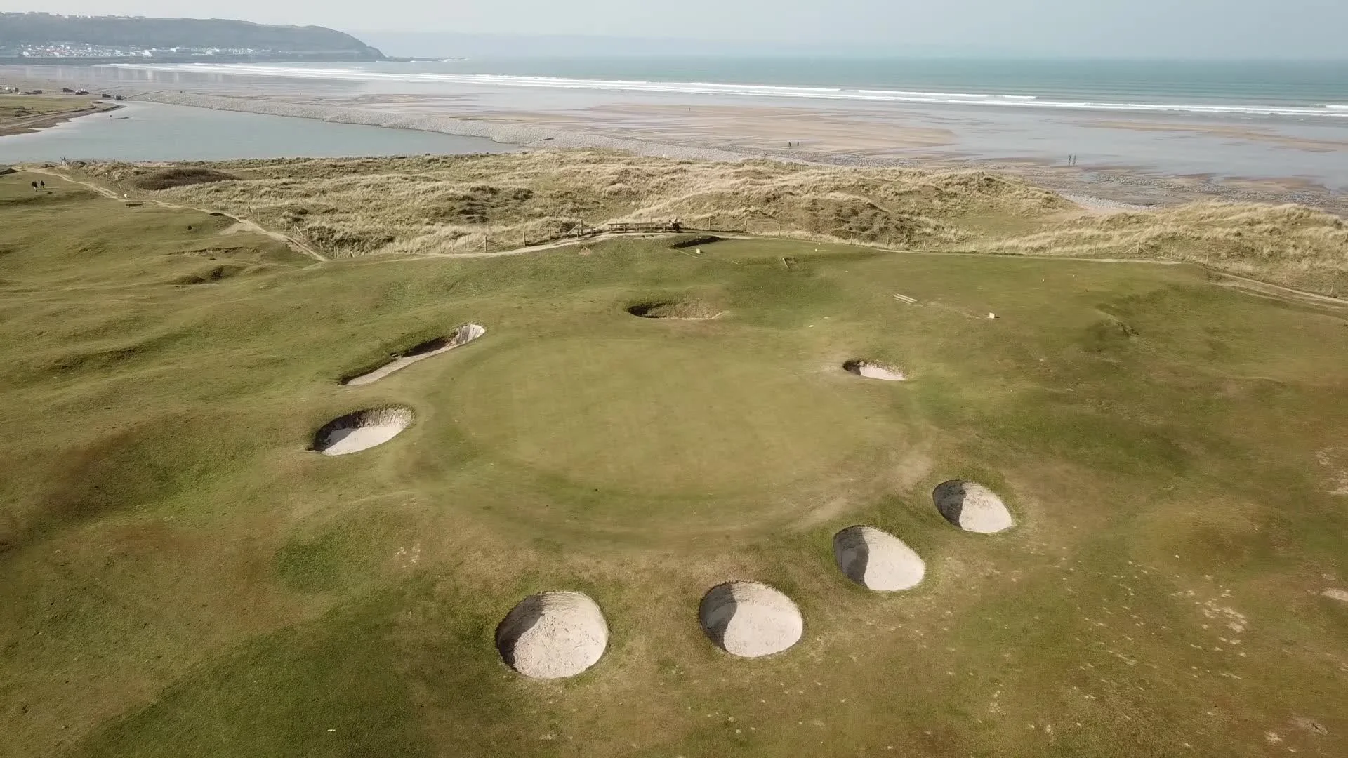 Aerial view of a green ringed by pot bunkers at Royal North Devon, with dunes, an estuary and a long sandy beach beyond