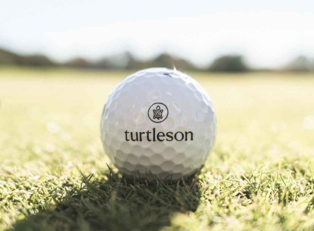 A white Turtleson golf ball with the turtle logo sitting on the grass, with the fairway blurred behind
